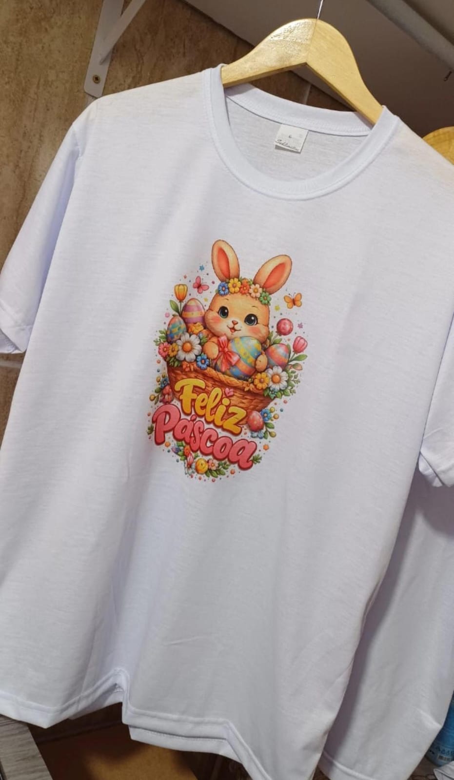 Camiseta branca com estampa de coelhinho em uma cesta de ovos coloridos e flores e o texto Feliz Páscoa