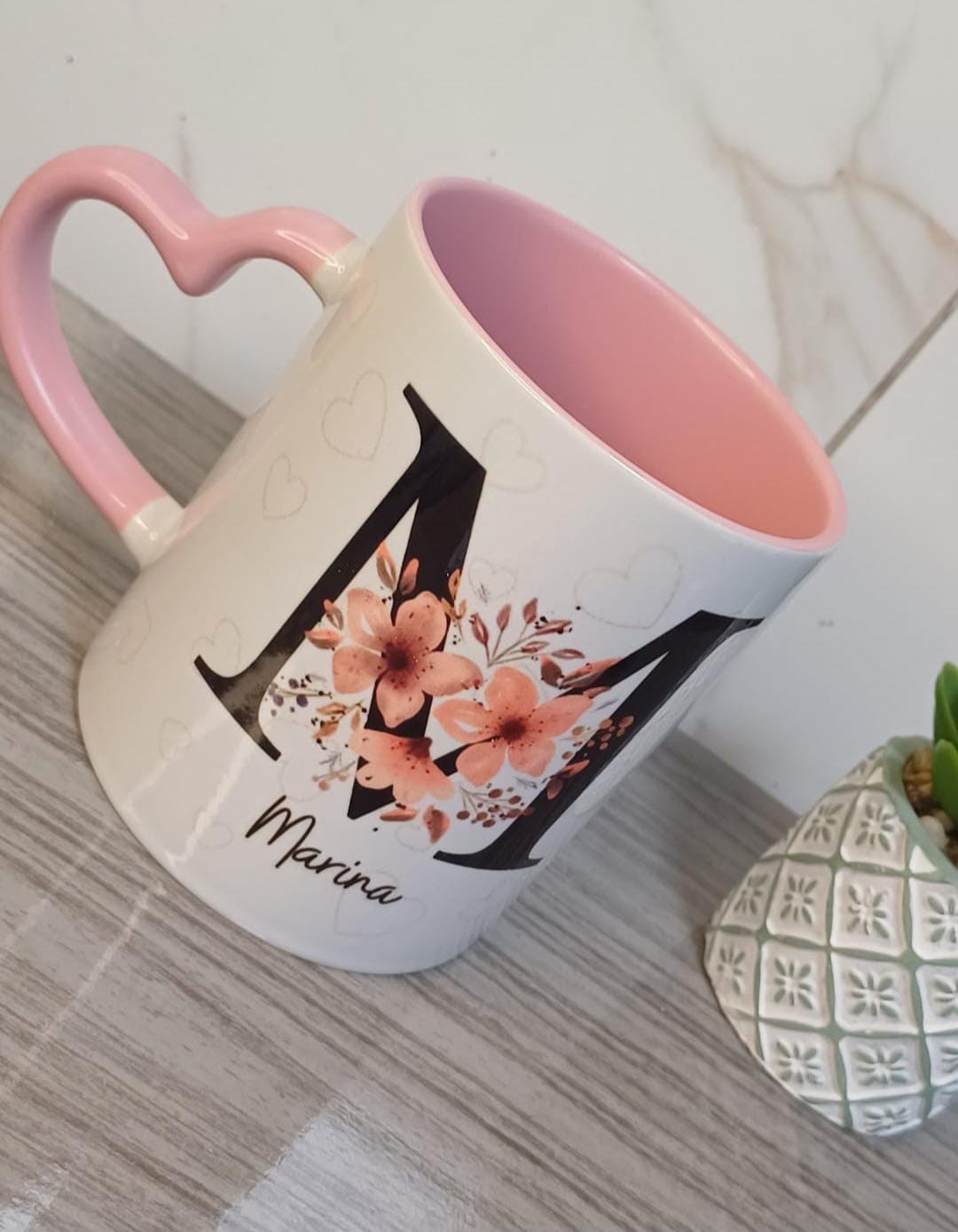 Caneca branca com interior e alça de coração rosa, letra M grande com flores e o nome Marina