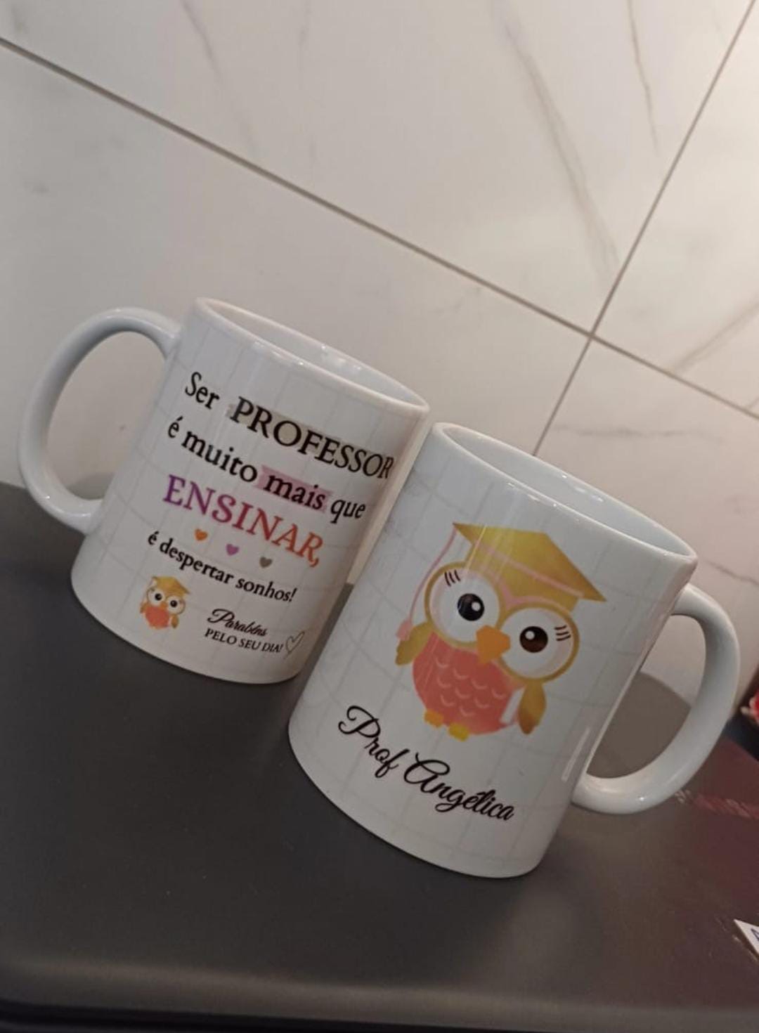 Duas canecas brancas com coruja de formatura, a frase Ser professor é muito mais que ensinar e o nome Prof Angélica