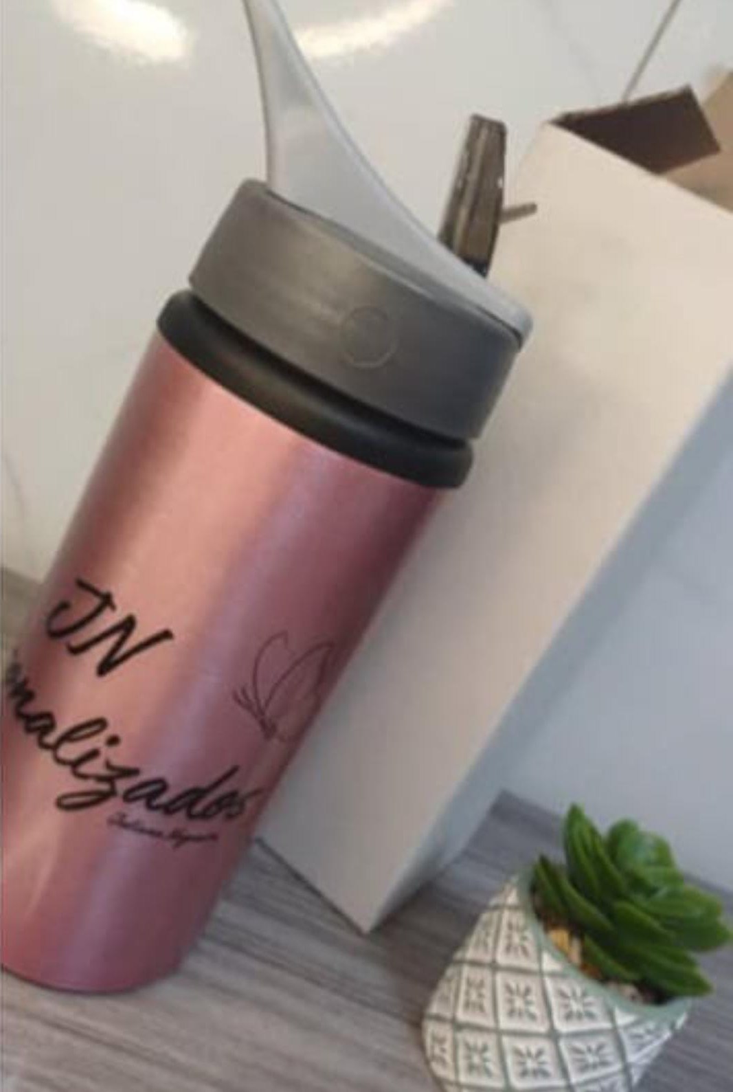 Squeeze rosa metalizado com tampa cinza e bico de canudo, com nome em letra cursiva preta e desenho de borboleta