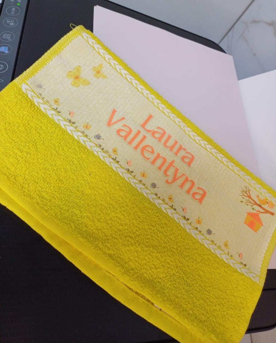 Toalhinha amarela com faixa estampada de borboletas, flores e casinha de passarinho e o nome Laura Vallentyna